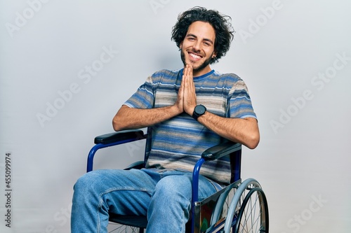 Handsome hispanic man sitting on wheelchair praying with hands together asking for forgiveness smiling confident.