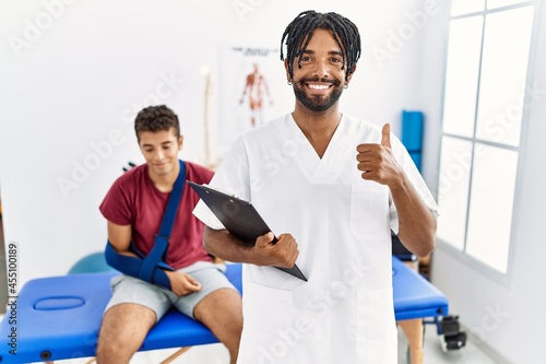 Young hispanic man working at pain recovery clinic with a man with broken arm doing happy thumbs up gesture with hand. approving expression looking at the camera showing success.