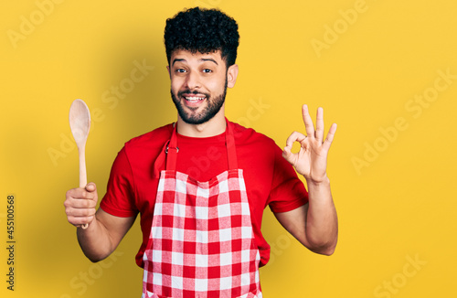 Young arab man with beard wearing baker uniform holding wooden spoon celebrating crazy and amazed for success with open eyes screaming excited.