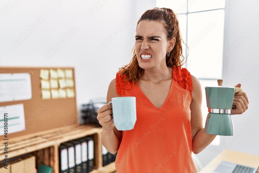 Young redhead woman holding french coffee maker at the office angry and mad screaming frustrated and furious, shouting with anger. rage and aggressive concept. Young redhead woman holding french coffee maker at the office angry and mad screaming frustrated and furious, shouting with anger. rage and aggressive concept.