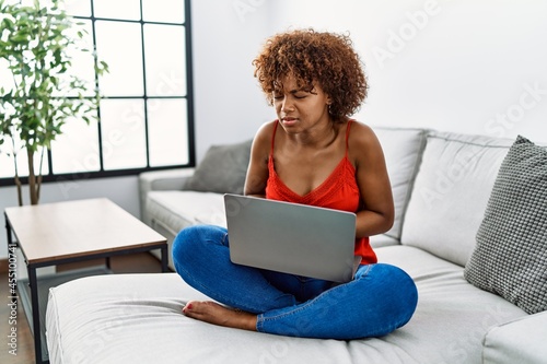 Young african american woman sitting on the sofa at home using laptop with hand on stomach because nausea, painful disease feeling unwell. ache concept.