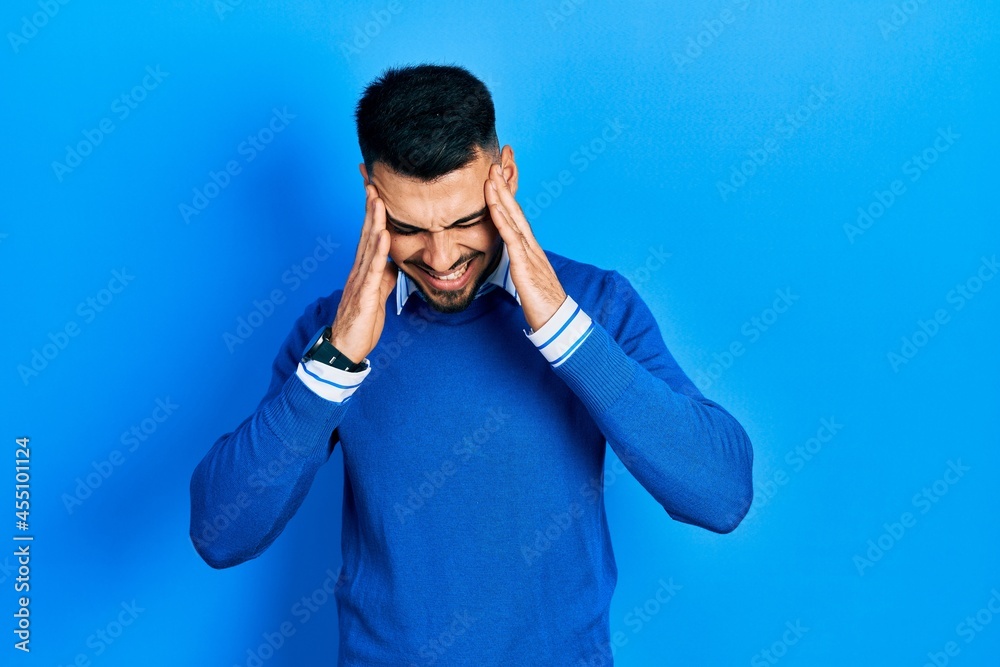 Young hispanic man with beard wearing casual blue sweater with hand on head, headache because stress. suffering migraine. Young hispanic man with beard wearing casual blue sweater with hand on head, headache because stress. suffering migraine.