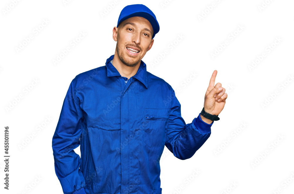 Bald man with beard wearing builder jumpsuit uniform with a big smile on face, pointing with hand finger to the side looking at the camera.