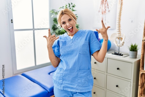 Beautiful blonde physiotherapist woman working at pain recovery clinic shouting with crazy expression doing rock symbol with hands up. music star. heavy concept.
