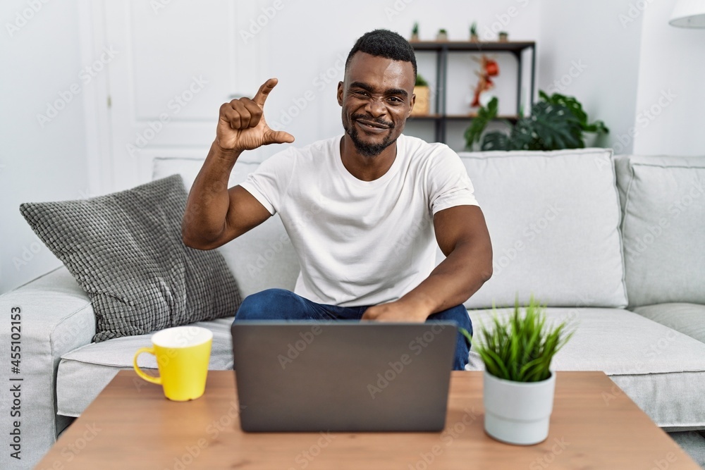 Young african man using laptop at home smiling and confident gesturing with hand doing small size sign with fingers looking and the camera. measure concept.