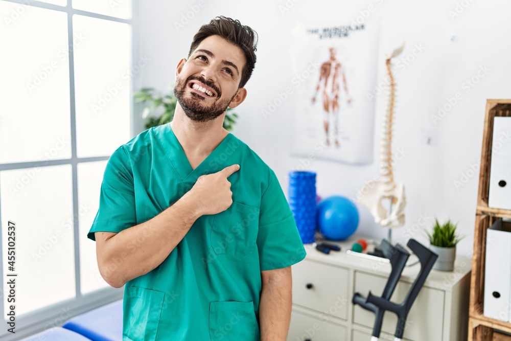 Young man with beard working at pain recovery clinic cheerful with a smile of face pointing with hand and finger up to the side with happy and natural expression on face Young man with beard working at pain recovery clinic cheerful with a smile of face pointing with hand and finger up to the side with happy and natural expression on face