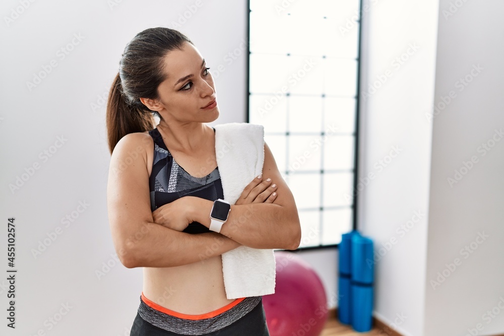Young brunette woman wearing sportswear and towel at the gym looking to the side with arms crossed convinced and confident