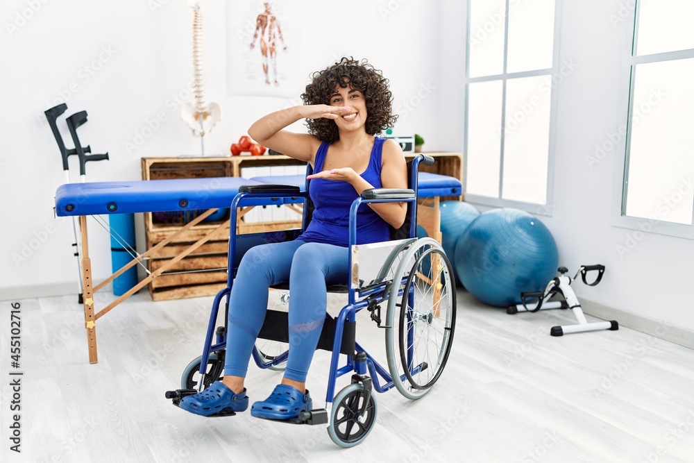 Fototapeta premium Young middle eastern woman sitting on wheelchair at physiotherapy clinic gesturing with hands showing big and large size sign, measure symbol. smiling looking at the camera. measuring concept.