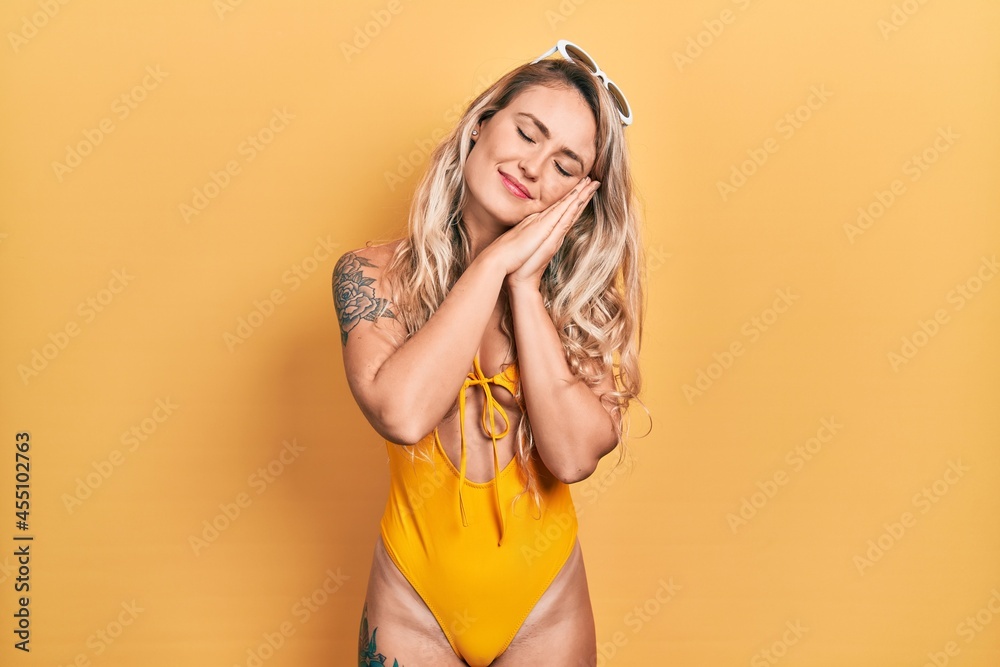 Beautiful young blonde woman wearing swimsuit and sunglasses sleeping tired dreaming and posing with hands together while smiling with closed eyes. Beautiful young blonde woman wearing swimsuit and sunglasses sleeping tired dreaming and posing with hands together while smiling with closed eyes.