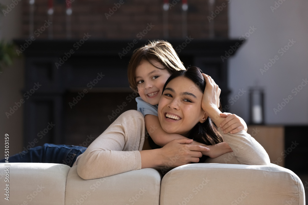 Cheerful small cute kid boy cuddling beautiful vietnamese asian mother or female nanny, resting together on huge cozy couch, happy different generations mixed race family spending weekend together. Cheerful small cute kid boy cuddling beautiful vietnamese asian mother or female nanny, resting together on huge cozy couch, happy different generations mixed race family spending weekend together.