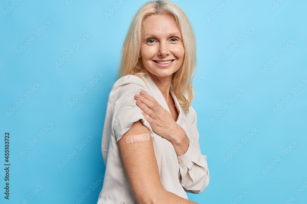 Sideways shot of blonde forty years old woman approves immunization campaign gets inoculation from covid 19 virus feels protected isolated over blue background. Vaccinated middle aged European lady - obrazy, fototapety, plakaty 