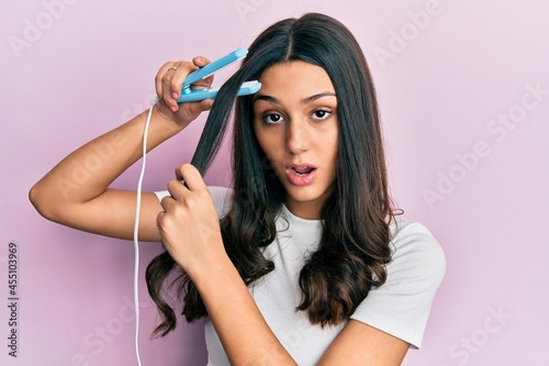 Young hispanic woman holding hair straightener in shock face, looking skeptical and sarcastic, surprised with open mouth