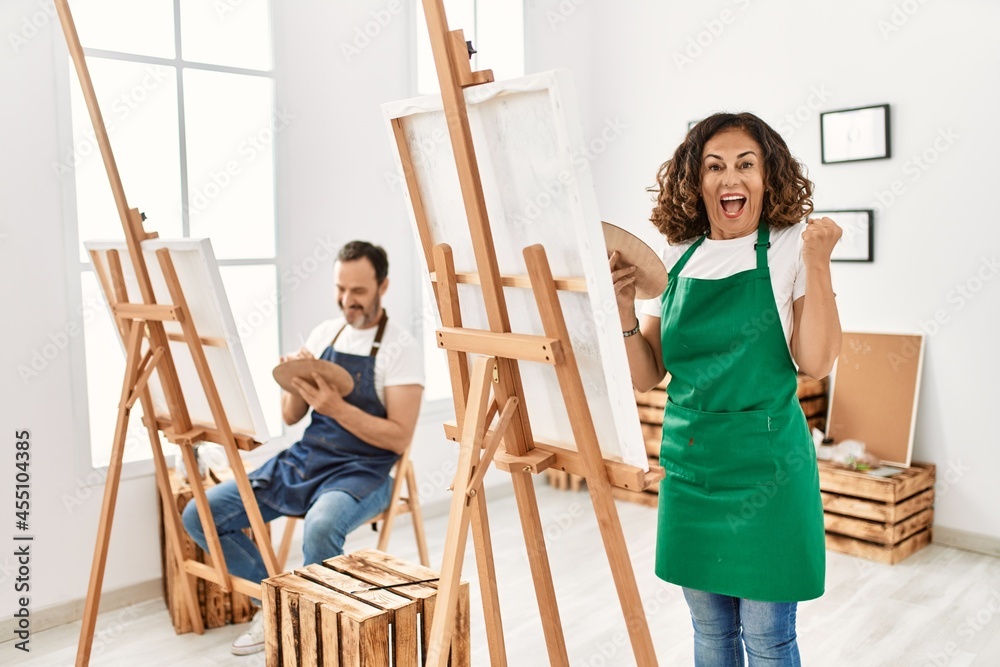 Hispanic middle age woman and mature man at art studio screaming proud, celebrating victory and success very excited with raised arms