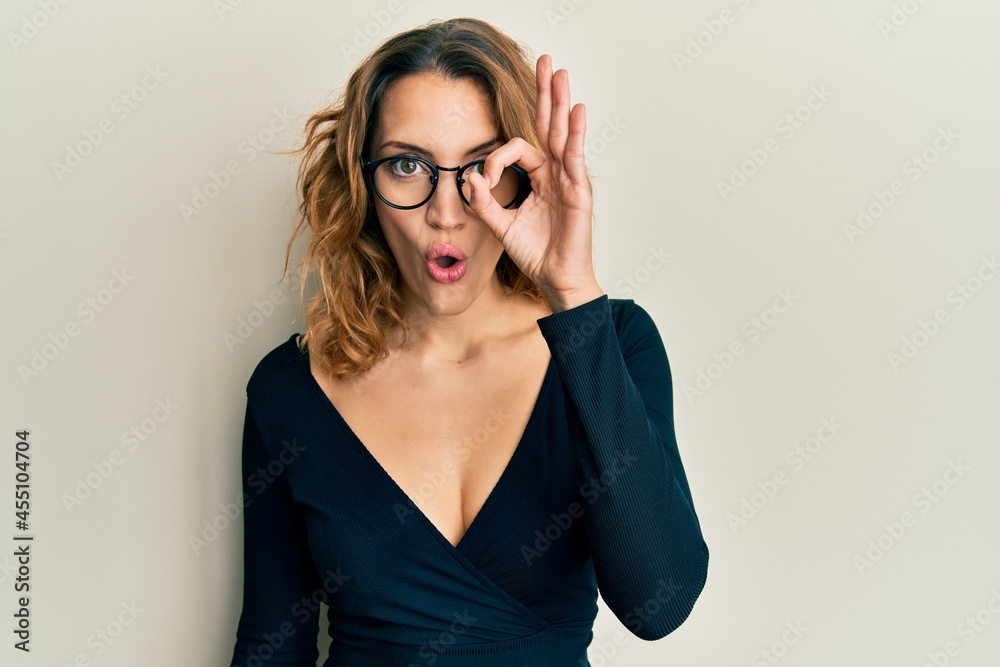 Young caucasian woman wearing business shirt and glasses doing ok gesture shocked with surprised face, eye looking through fingers. unbelieving expression.