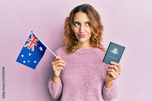 Young caucasian woman holding australian flag and passport smiling looking to the side and staring away thinking.