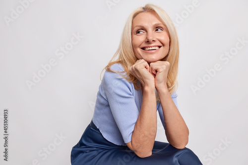 Indoor shot of dreamy middle aged blonde woman keeps hands under chin recalls pleasant memory in mind smiles broadly sits against grey background blank empty space for your advertisement or text
