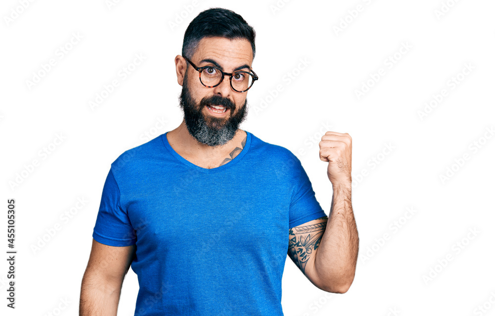 Hispanic man with beard wearing casual t shirt and glasses smiling with happy face looking and pointing to the side with thumb up. Hispanic man with beard wearing casual t shirt and glasses smiling with happy face looking and pointing to the side with thumb up.