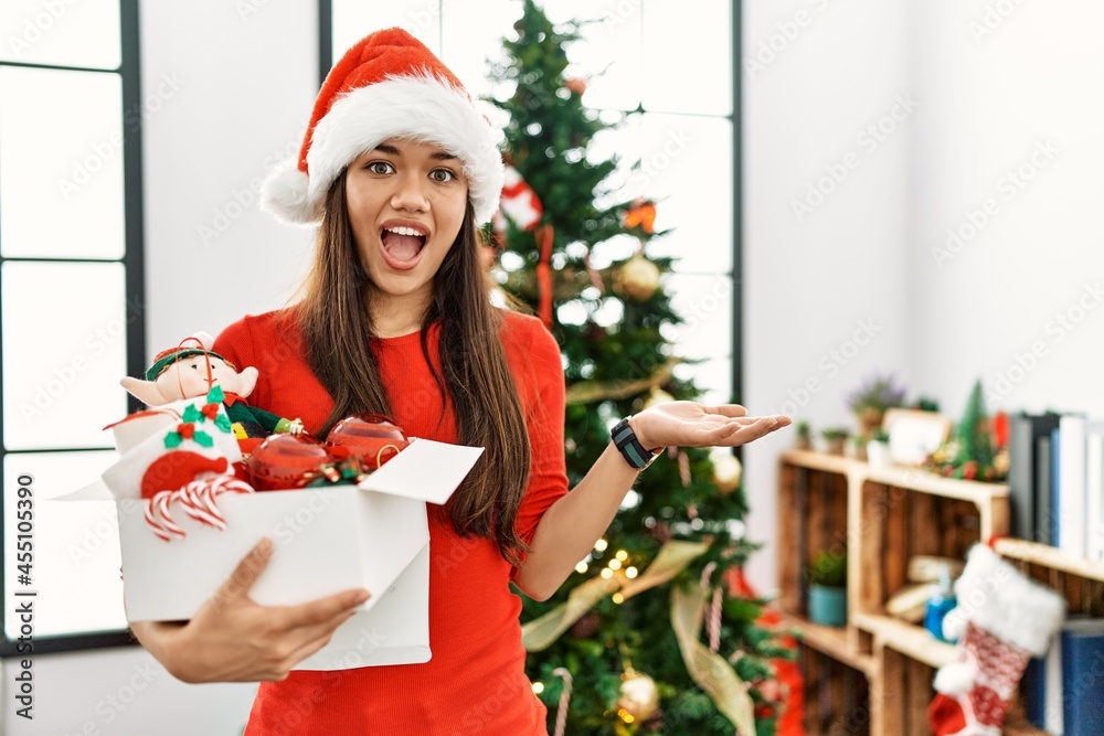 Young brunette woman standing by christmas tree holding decoration pointing aside with hands open palms showing copy space, presenting advertisement smiling excited happy