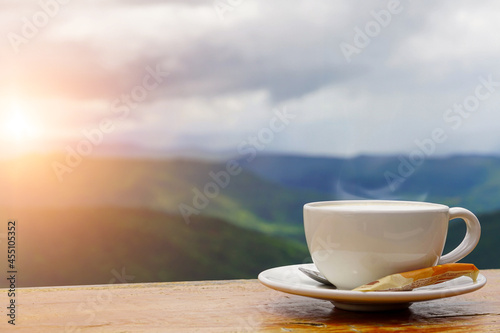 A white cup of hot espresso coffee mugs placed on a wooden floor with morning fog and mountains with sunlight background,coffee morning