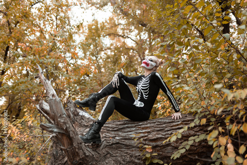 A girl with a scary make-up in a skeleton costume, a mask with rhinestones in a gloomy autumn forest. Sits in a tree. Portrait. Halloween, autumn holiday concept 