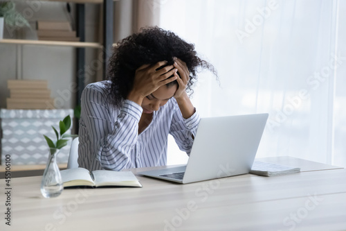 Professional burnout. Depressed upset mixed race female sit at workplace by computer hug head feel unmotivated sick lack of sleep. Desperate young black woman having problems at work missed deadline
