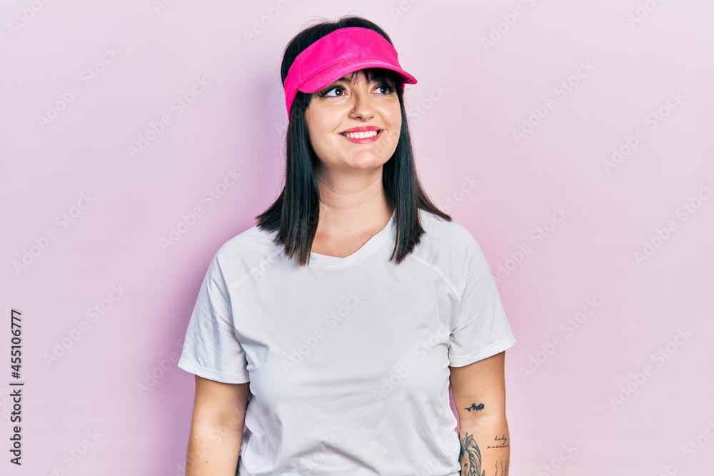 Young hispanic woman wearing sportswear and sun visor cap looking away to side with smile on face, natural expression. laughing confident. Young hispanic woman wearing sportswear and sun visor cap looking away to side with smile on face, natural expression. laughing confident.