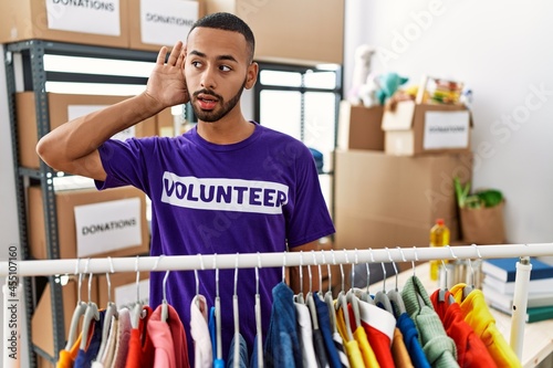 African american man wearing volunteer t shirt at donations stand smiling with hand over ear listening an hearing to rumor or gossip. deafness concept.