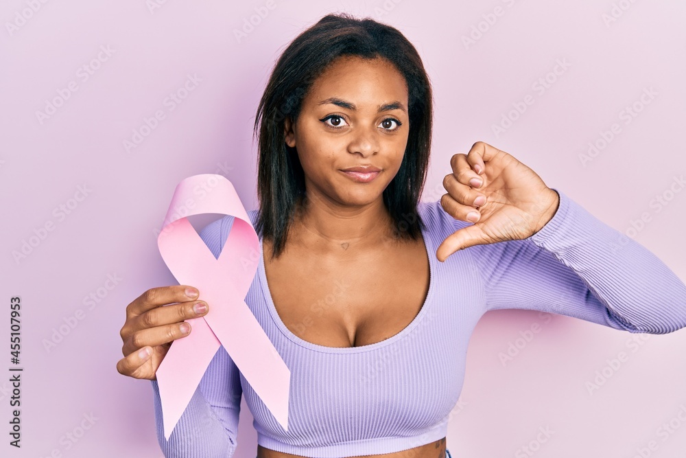 Young african american girl holding pink cancer ribbon with angry face, negative sign showing dislike with thumbs down, rejection concept Young african american girl holding pink cancer ribbon with angry face, negative sign showing dislike with thumbs down, rejection concept