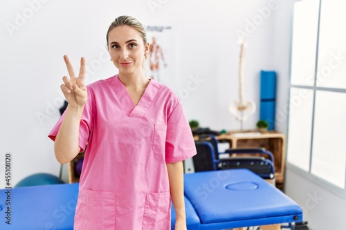 Young blonde woman working at pain recovery clinic showing and pointing up with fingers number two while smiling confident and happy.