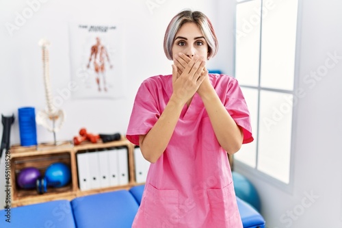 Young nurse woman working at pain recovery clinic shocked covering mouth with hands for mistake. secret concept.