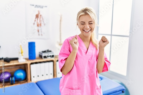 Young caucasian woman working at pain recovery clinic excited for success with arms raised and eyes closed celebrating victory smiling. winner concept.