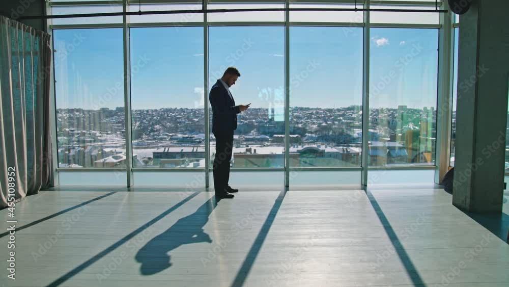 Businessman with a phone indoors. Young entrepreneur in suit standing in spacious room near large windows and uses mobile phone. Businessman with a phone indoors. Young entrepreneur in suit standing in spacious room near large windows and uses mobile phone.