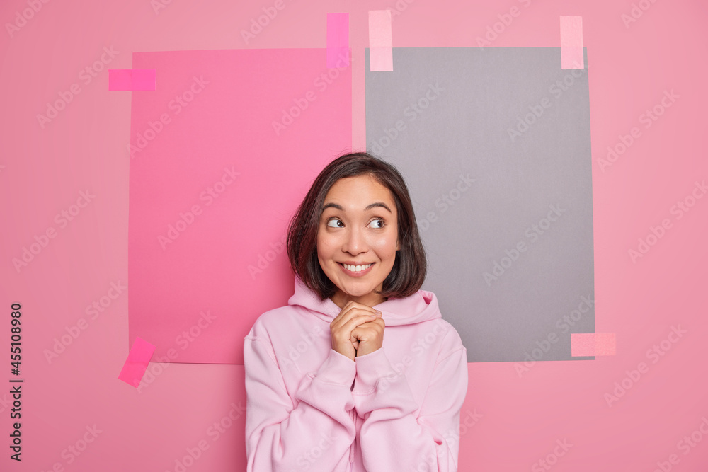 Horizontal shot of satisfied Asian woman keeps hands together smiles pleasantly wears hoodie thinks about something pleasant poses against pink background with two plastered sheets of paper. Horizontal shot of satisfied Asian woman keeps hands together smiles pleasantly wears hoodie thinks about something pleasant poses against pink background with two plastered sheets of paper.