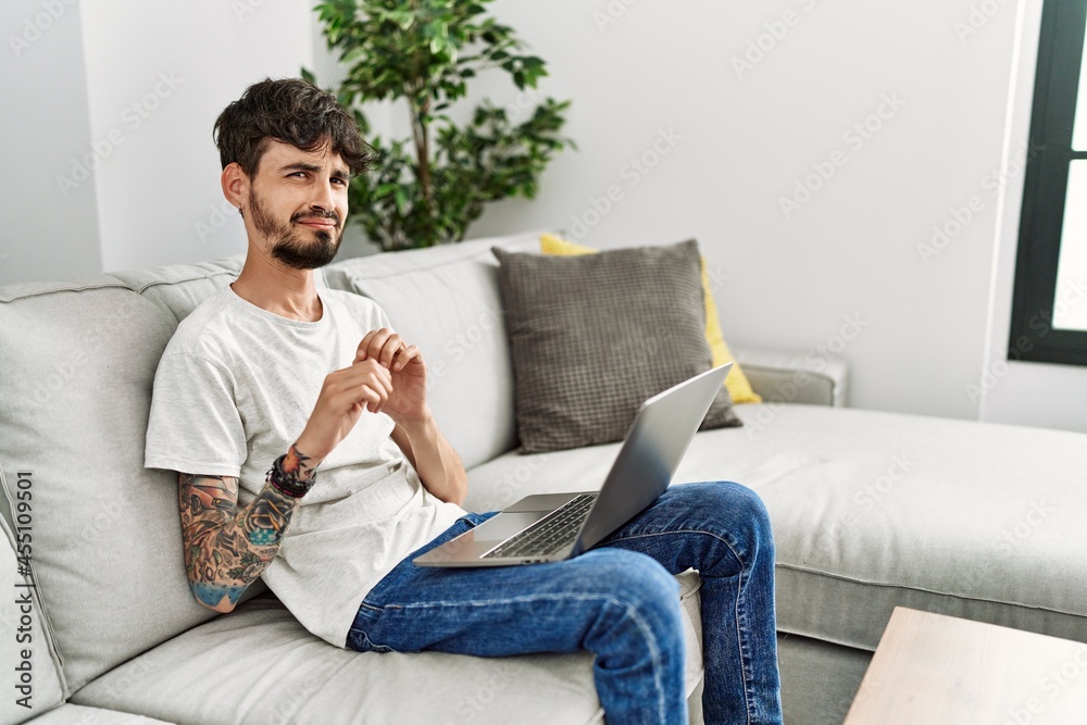 Hispanic man with beard sitting on the sofa disgusted expression, displeased and fearful doing disgust face because aversion reaction. with hands raised