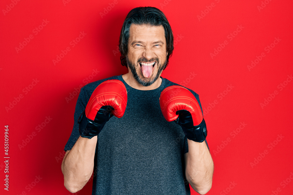 Middle age caucasian man using boxing gloves sticking tongue out happy with funny expression.