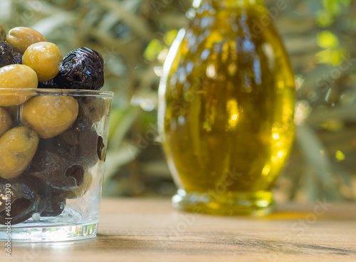 Black and green olives in a transparent bowl with olive oil and olive tree in the background