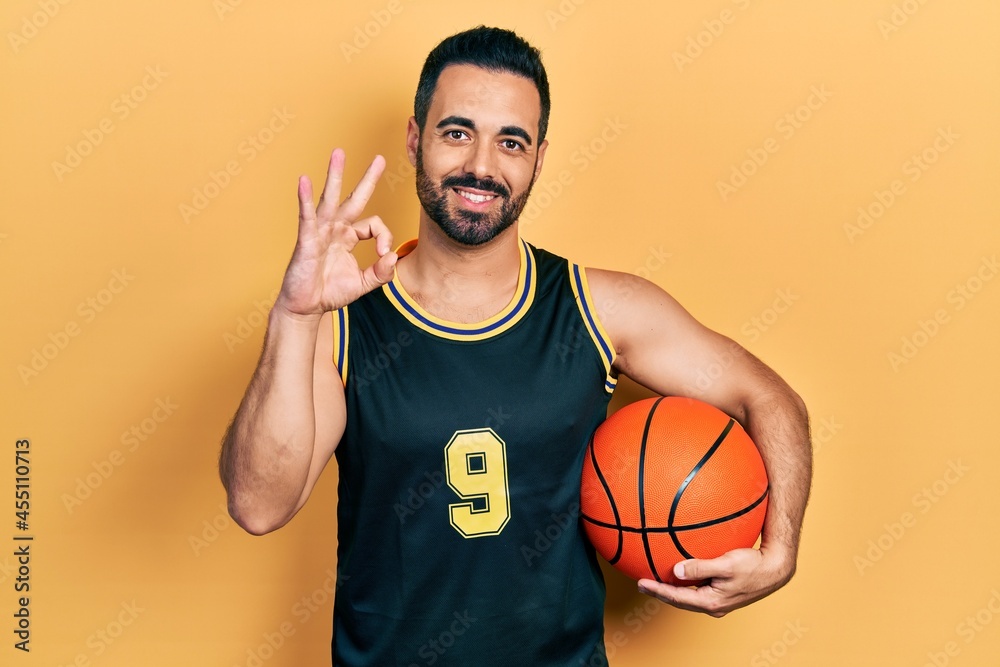 Handsome hispanic man with beard holding basketball ball doing ok sign with fingers, smiling friendly gesturing excellent symbol