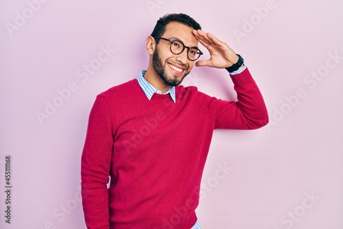 Hispanic man with beard wearing business shirt and glasses very happy and smiling looking far away with hand over head. searching concept.