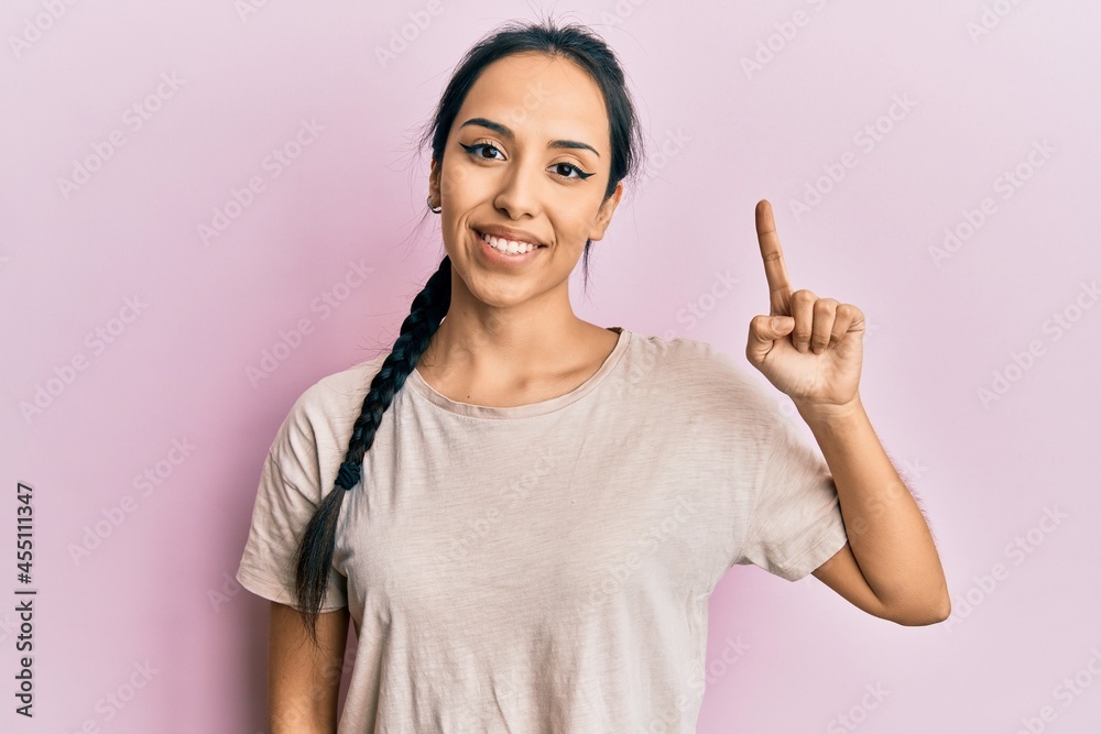 Young hispanic girl wearing casual white t shirt smiling with an idea or question pointing finger up with happy face, number one Young hispanic girl wearing casual white t shirt smiling with an idea or question pointing finger up with happy face, number one