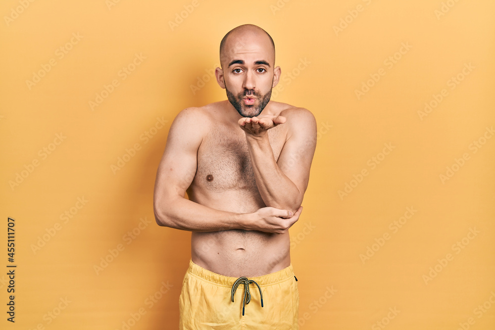 Young bald man wearing swimwear looking at the camera blowing a kiss with hand on air being lovely and sexy. love expression. Young bald man wearing swimwear looking at the camera blowing a kiss with hand on air being lovely and sexy. love expression.
