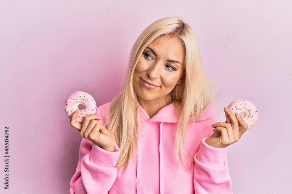 Young blonde woman holding tasty pink doughnuts over eyes smiling looking to the side and staring away thinking. Young blonde woman holding tasty pink doughnuts over eyes smiling looking to the side and staring away thinking.
