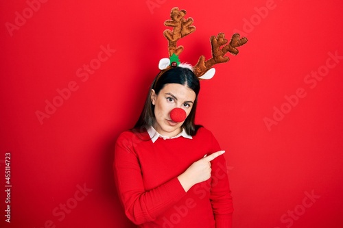 Young hispanic woman wearing deer christmas hat and red nose pointing with hand finger to the side showing advertisement, serious and calm face