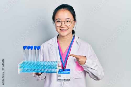 Young chinese girl wearing scientist uniform holding test tube smiling happy pointing with hand and finger