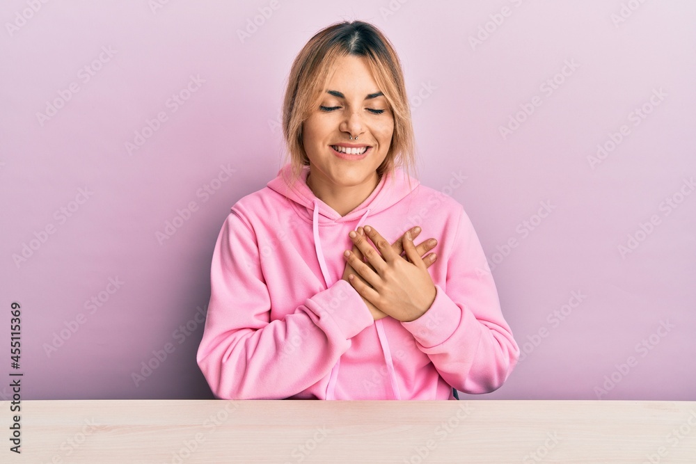 Young caucasian woman wearing casual clothes sitting on the table smiling with hands on chest with closed eyes and grateful gesture on face. health concept. Young caucasian woman wearing casual clothes sitting on the table smiling with hands on chest with closed eyes and grateful gesture on face. health concept.