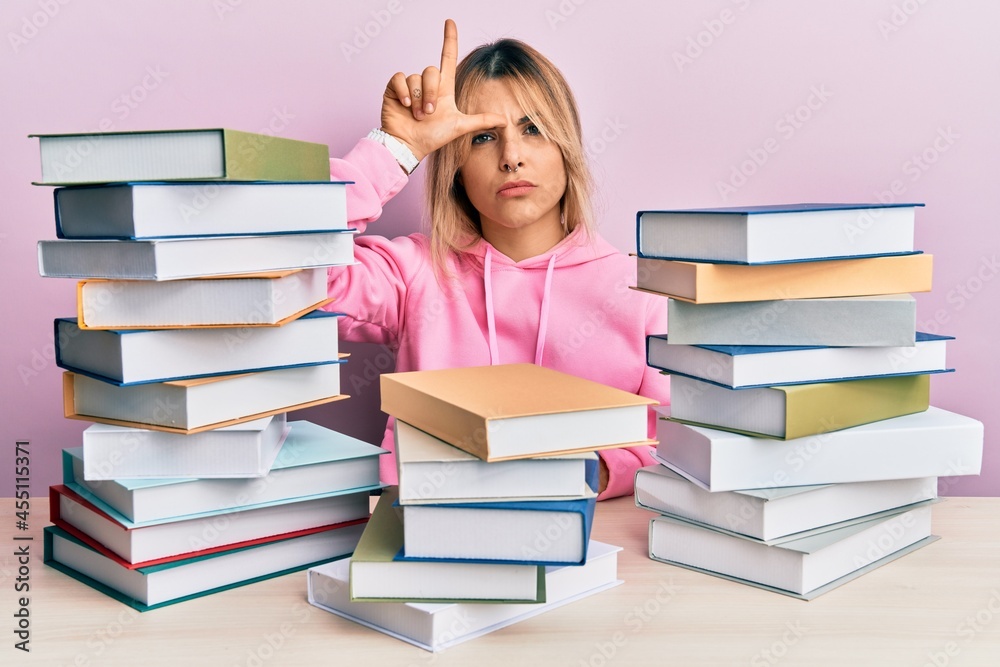 Young caucasian woman sitting on the table with books making fun of people with fingers on forehead doing loser gesture mocking and insulting. Young caucasian woman sitting on the table with books making fun of people with fingers on forehead doing loser gesture mocking and insulting.