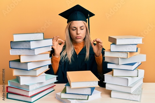 Young caucasian woman wearing graduation ceremony robe sitting on the table relaxed and smiling with eyes closed doing meditation gesture with fingers. yoga concept.