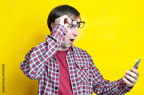 handsome surprised and excited man with open mouth in red checkered shirt taking off glasses on forehead and looking at smartphone on yellow background with copy space