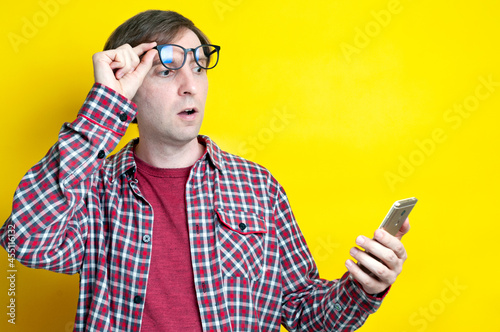 handsome amazed man with open mouth in red checkered shirt taking off glasses and looking at smartphone on yellow background with copy space