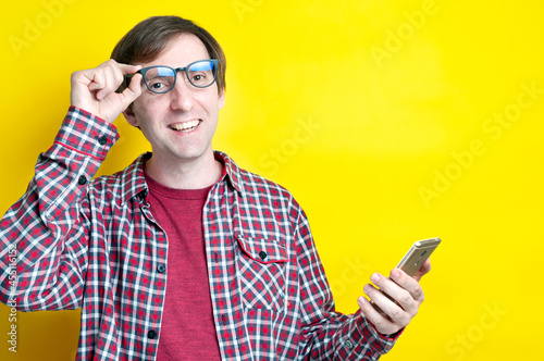 handsome cheerful smiling man in red t shirt putting on glasses, holding smartphone and looking at camera on  yellow background with copy space