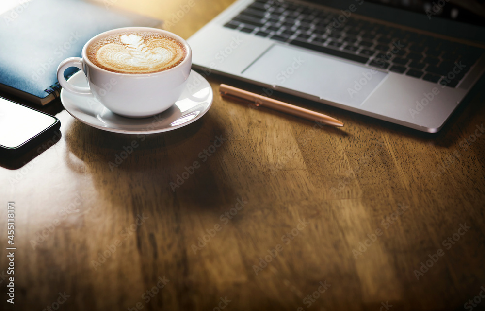 Focus on latte art coffee in white cup with computer laptop, mobile phone and blue note book on wooden table Focus on latte art coffee in white cup with computer laptop, mobile phone and blue note book on wooden table
